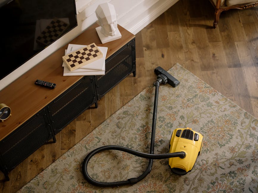 Close-up of a vacuum cleaner head actively cleaning a patterned carpet with floral motifs in a residential living room. The carpet features a mix of beige, light green, and orange tones, and appears t
