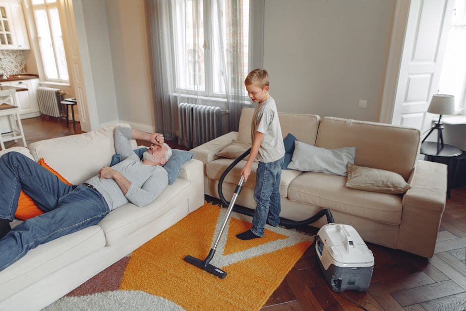 A person is vacuuming a patterned area rug on a wooden floor in a living room. The individual, wearing casual clothing and sneakers, is holding a handheld vacuum cleaner with a long wand attachment. T