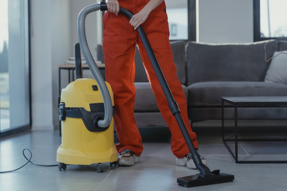 A professional cleaner wearing orange overalls vacuuming a modern living room with a yellow industrial vacuum cleaner. The room features a tiled floor, a dark gray fabric sofa with light-colored cushi