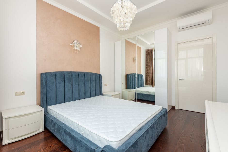A modern bedroom featuring a low-profile beige upholstered bed with a purple mattress, situated on a dark blue carpet. The room has white walls and built-in storage cabinets with a sleek, handle-free 