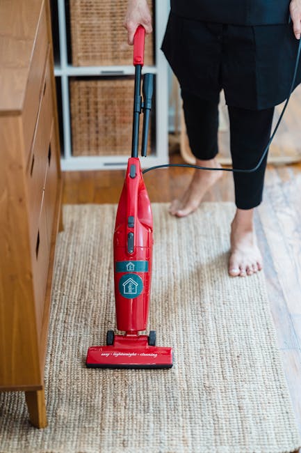 A person wearing beige trousers and white sneakers is vacuuming a patterned area rug with a yellow and black vacuum cleaner. The rug features a detailed floral motif in shades of beige, green, and lig
